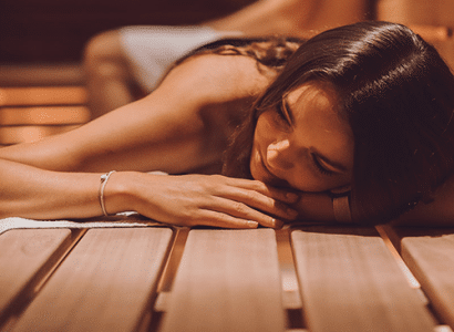Eine junge Frau ruht sich aus, mit dem Kopf auf den Armen liegend, in einer holzverkleideten Sauna der Therme Wien.