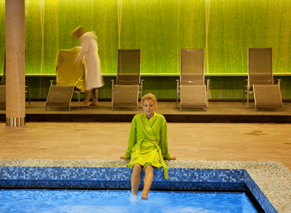 Eine Frau in einem gruenen Bademantel sitzt entspannt mit geschlossenen Augen am Rande eines blauen, gefliesten Pools in einem Innenbereich. Im Hintergrund ist eine Wand mit einer grünen Mosaiktextur zu sehen, vor der mehrere Liegestühle aufgereiht sind. Eine weitere Person in einem weißen Bademantel ist unscharf zu sehen, wie sie sich gerade einen Liegestuhl herrichtet. Die Szene vermittelt eine ruhige und entspannende Atmosphaere in einem Wellnessbereich.