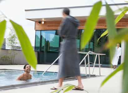 Ein Mann geniesst entspannt das Schwimmen in einem Außenpool der Therme Wien, waehrend eine Person in einem grauen Bademantel vorbeigeht und eine weitere Person in einem weissen Bademantel im Hintergrund steht, umgeben von gruenen Pflanzen, die teilweise die Szene rahmen.