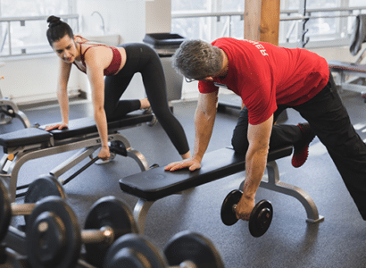 Frau beim Kurzhantelrudern mit ihrem Trainer. Dieser zeigt ihr die Uebung auf der Trainingsbank.