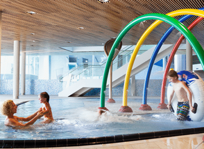 Eine erwachsene Frau spielt mit einem lachenden Kind in einem sprudelnden Pool unter bunten Wasserbogen in der Therme Wien. Ein weiteres Kind im Badeanzug eilt freudig ins Wasser, umgeben von einer zeitgenoessischen Architektur mit hellen Tageslichteinfall.