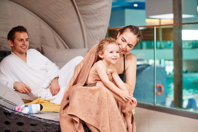 Familie sitzt in der Familienoase der Therme Wien. Frau wickelt ihr Kind in ein Handtuch, um es abzutrocknen. Mann liegt im Hintergrund und laechelt.