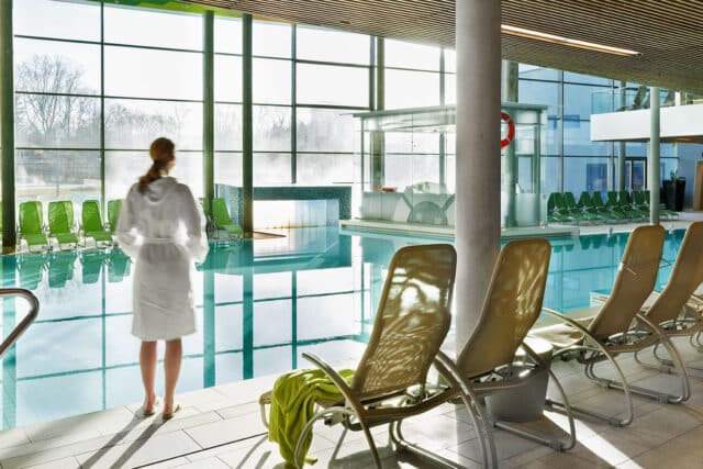 Frau im Bademantel steht am Beckenrand der Thermenlandschaft in der Therme Wien.