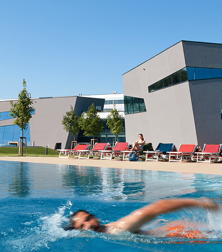 Mann krault im Sportbecken im Aussenbereich der Therme Wien. Es scheint die Sonne. Im Hintergrund sitzt eine Frau in der Liege und sonnt sich.