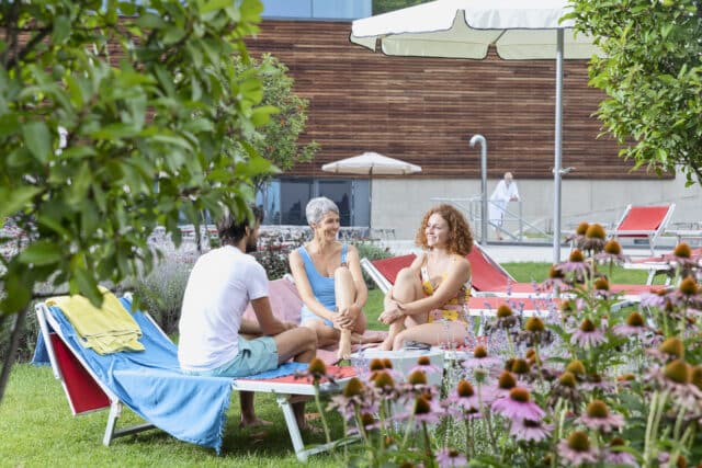 Drei Personen geniessen das schoene im Therme Wien Garten. Sie sitzen auf ihren Liegen und unterhalten sich.