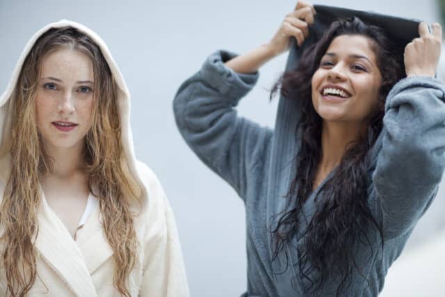 Zwei junge Frauen in Bademaenteln laecheln in die Kamera waehrend sie wellnessen.