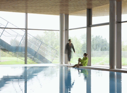 Fru sitzt mit gruenem Bademantel am Beckenrand der Therme Wien. Es naehert sich ein Mann in einem grauen Bademantel. Im Hintergrund ist der gruene Garten durch eine Glasfront zu sehen.