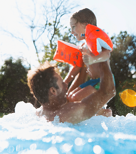 Vater und Sohn baden im Becken im Aussenbereich der Therme Wien. Der Vater hebt seinen Sohn aus dem Wasser. Der Sohn hat Schwimmfluegerl an. Es scheint die Sonne.